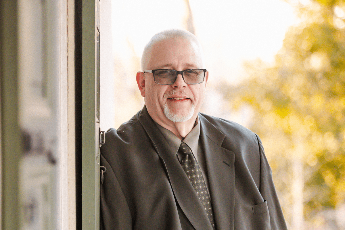 A man in glasses and a green suit posing on a porch on a sunny day.