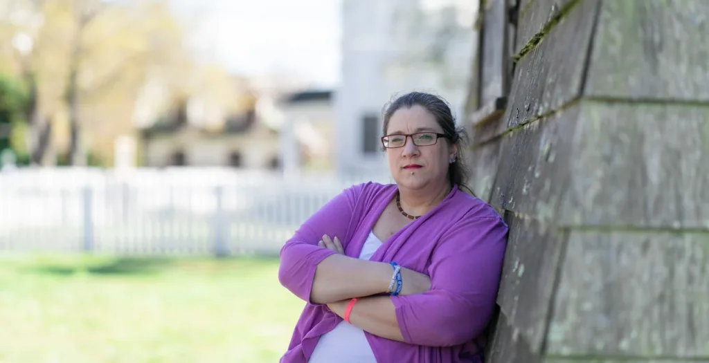 Ashley Kjarbo, plaintiff in Kjarbo v. NETU, IRS, leaning against a weathered wooden shingled house.